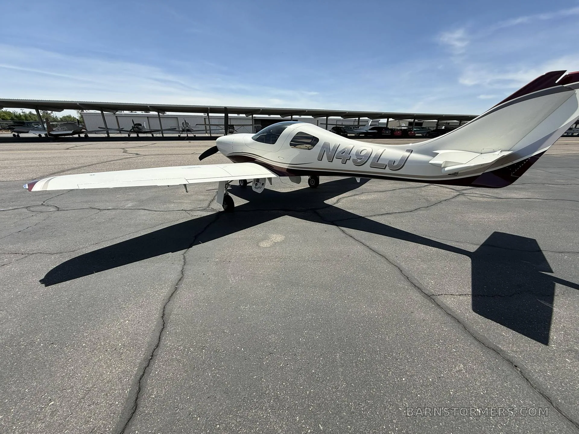 A rear three-quarter view of a white and maroon single-engine aircraft (N49LJ) parked on an asphalt apron with hangars in the background.
