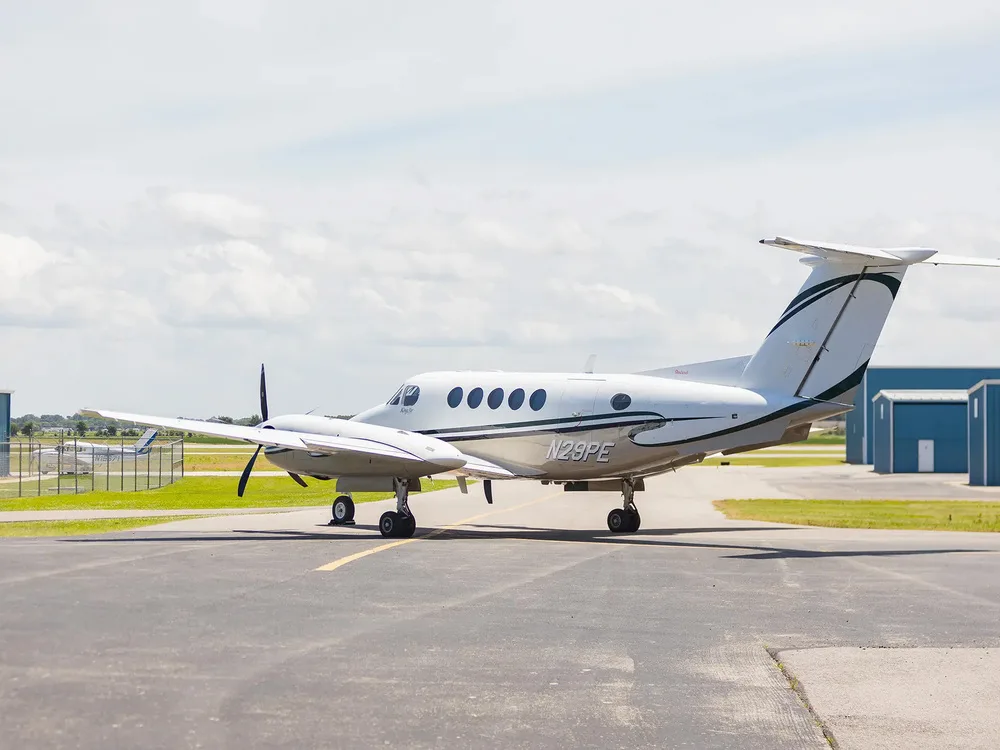A side view of the white Beechcraft King Air N29PE with dark green and black stripes parked on an airfield.