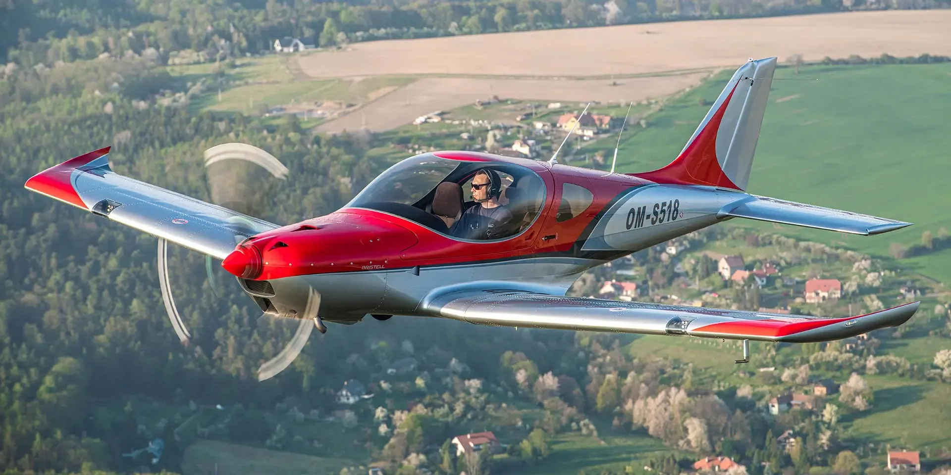 A red and silver Bristell light sport aircraft with a pilot flying over a green, residential landscape.