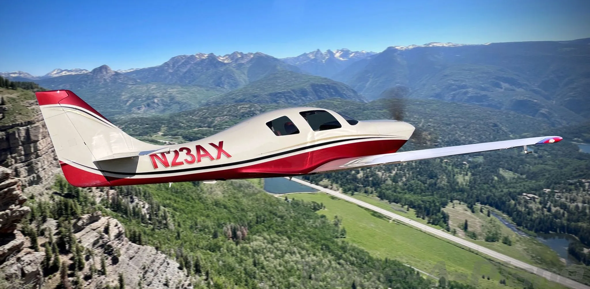 A white and red single-engine aircraft, N23AX, flies over a mountainous landscape with forests and a lake.
