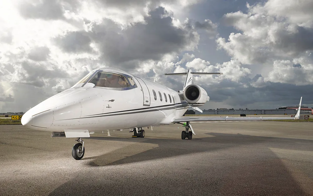 A white Learjet with black and grey stripes parked on a tarmac under a cloudy sky, viewed from the front-left.