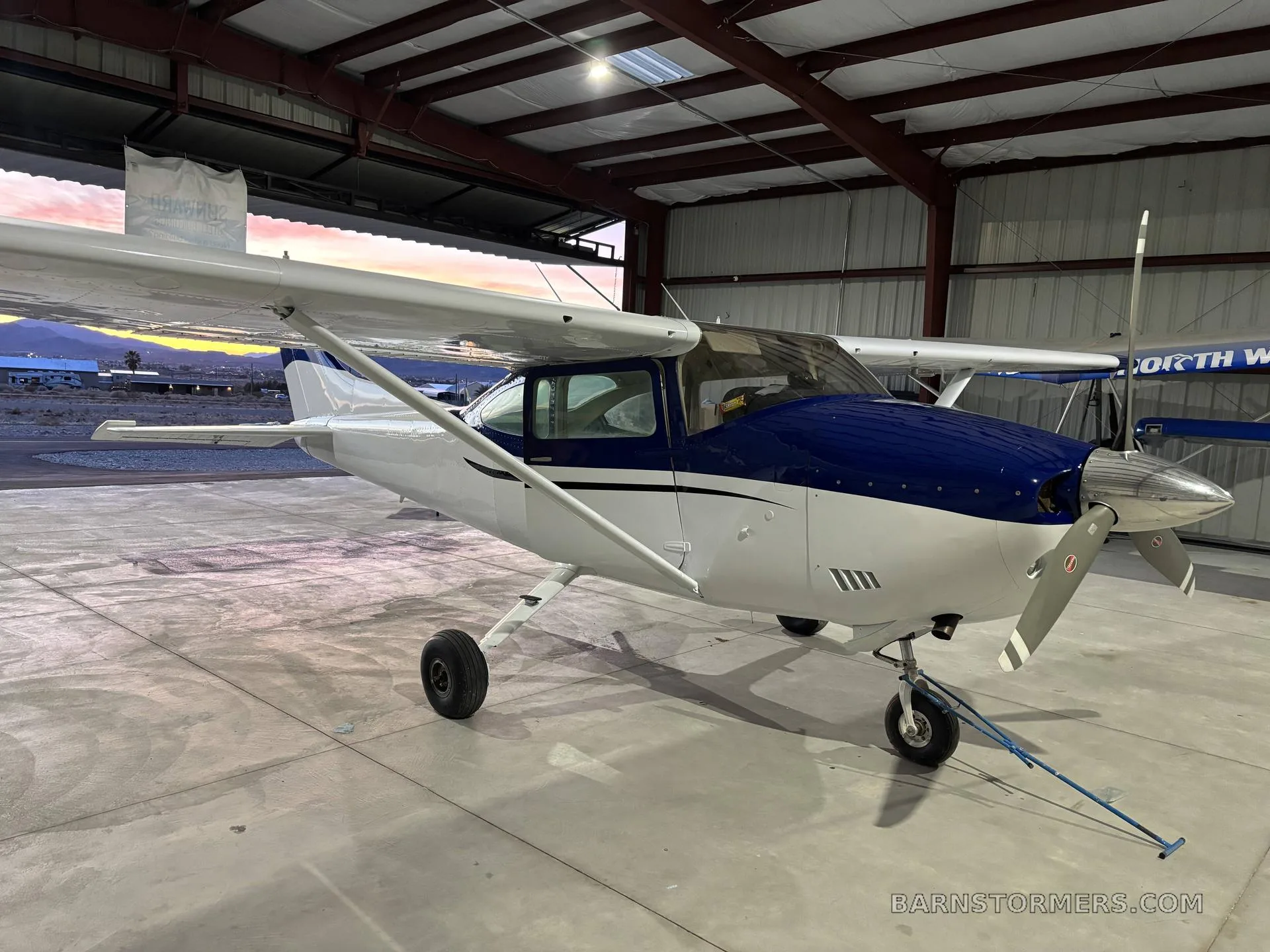 A blue and white single-engine propeller aircraft is parked inside a hangar, with another aircraft partially visible in the background.