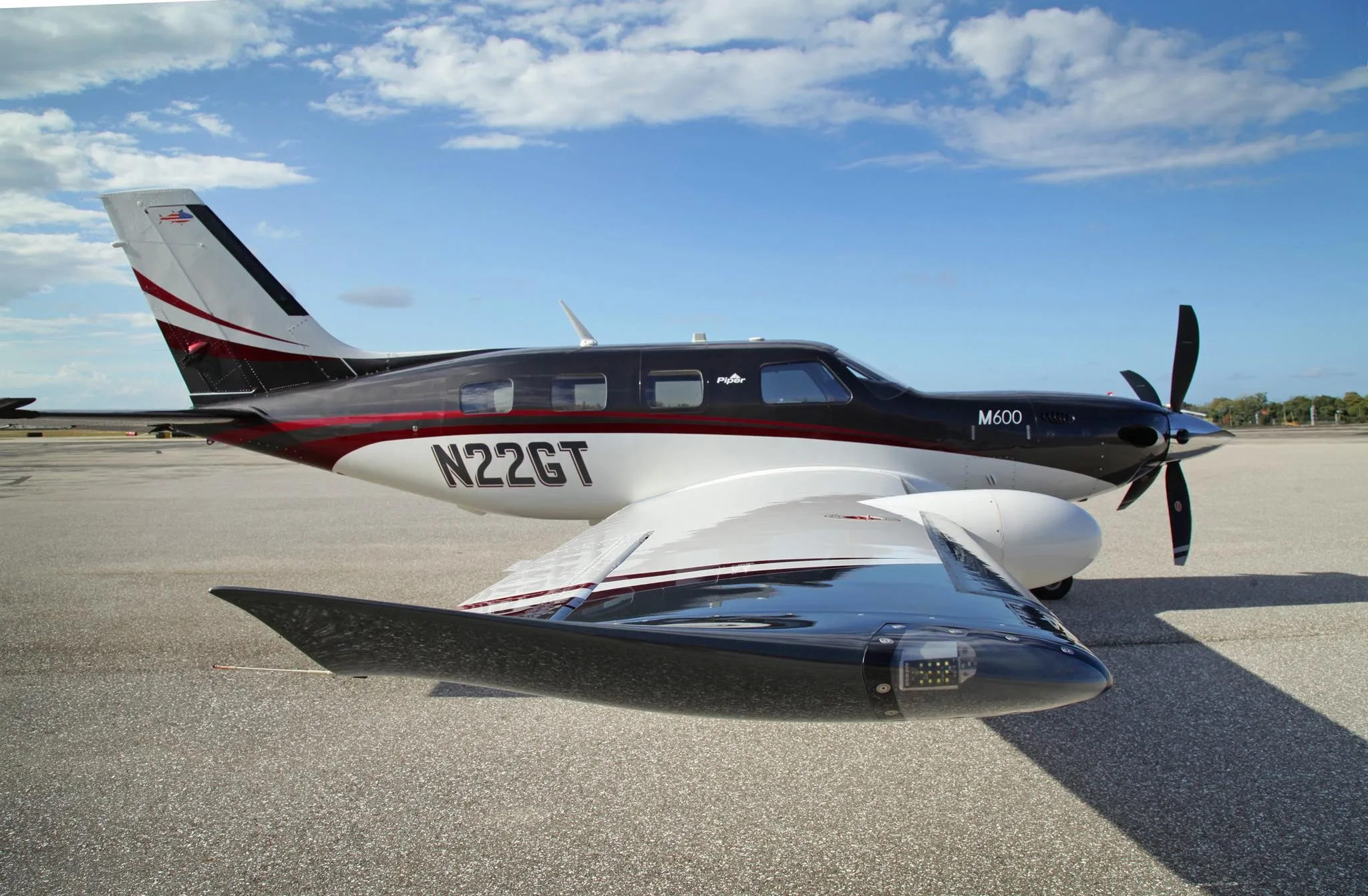 A white and black Piper M600 turboprop with red accents, parked on an airfield under a partly cloudy blue sky.
