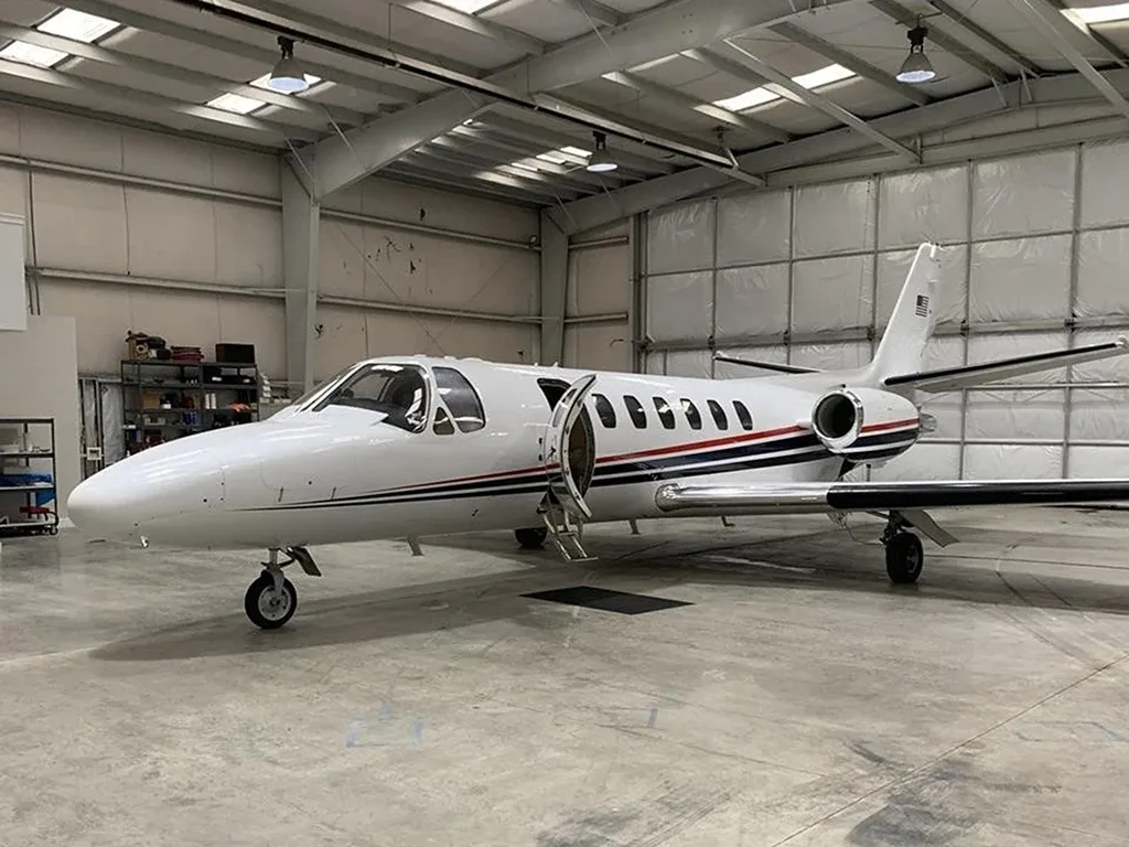 A white business jet with red and blue stripes parked inside a large hangar, with its main door open.