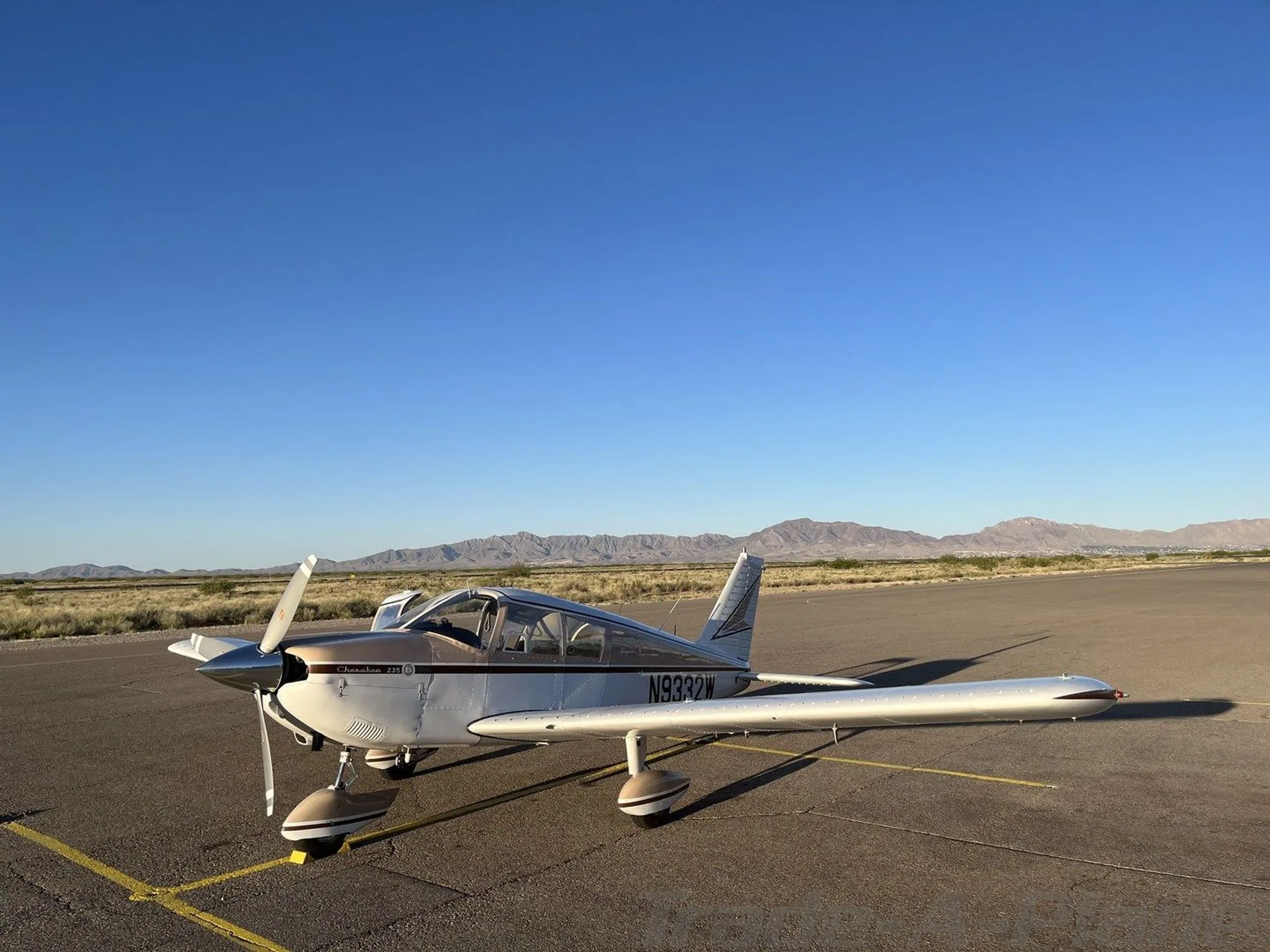 A Piper Cherokee 235, registration N9332W, in white and brown paint, parked on an airfield under a clear blue sky with mountains in the distance.
