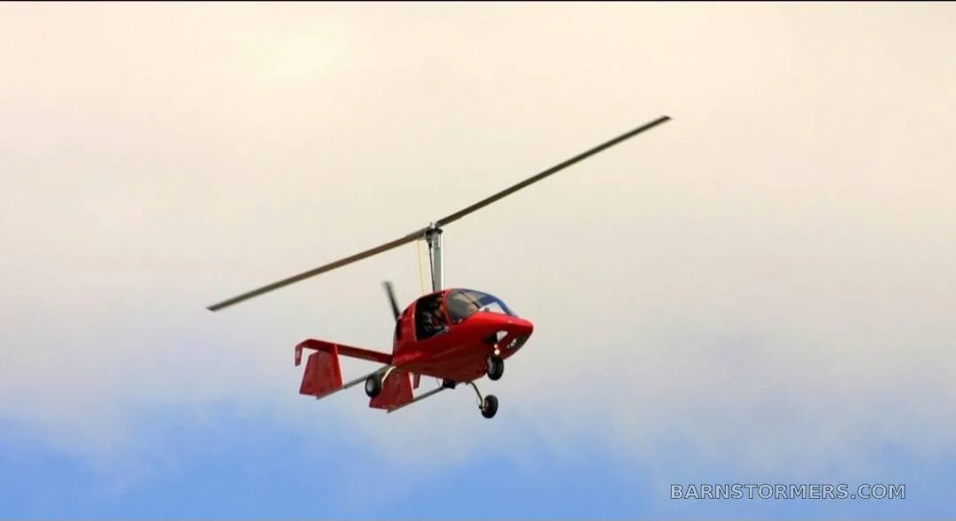 A red autogyro with a single main rotor and tail fins flies against a cloudy sky.