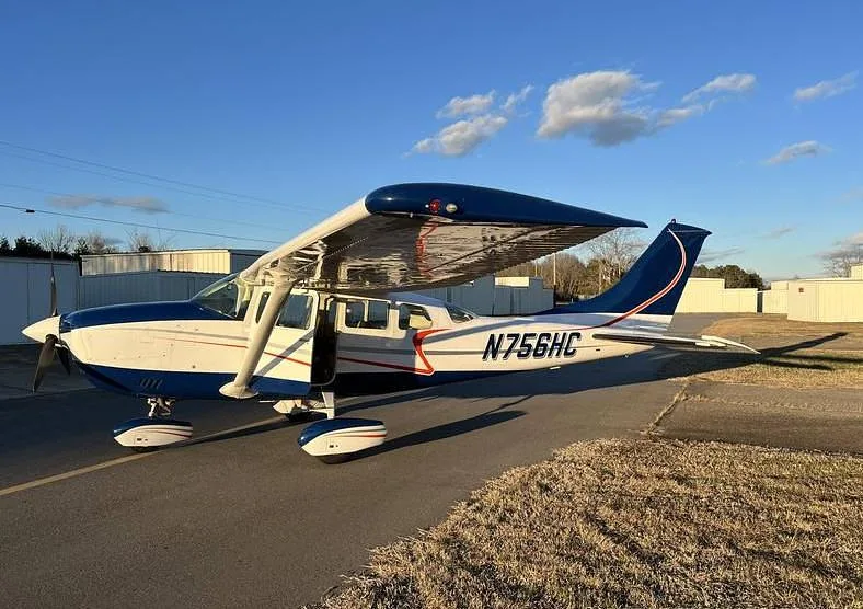A side view of a white and blue Cessna 182, N756HC, with its passenger door open, parked on an airfield next to dry grass.