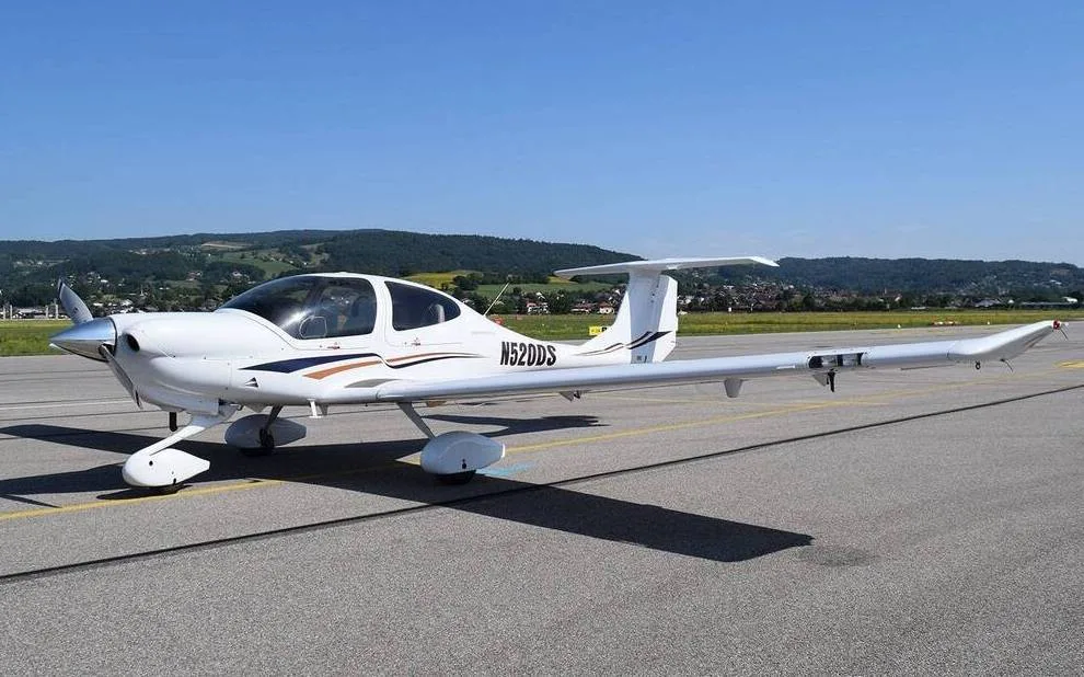 A white Diamond DA40 aircraft with brown and blue stripes parked on an airfield under a clear sky.