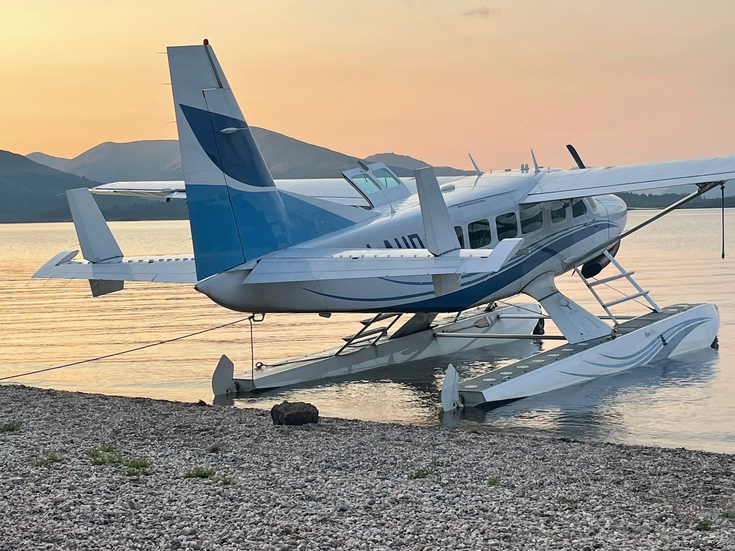 A white and blue floatplane, a Cessna Caravan, is tied to a rocky shore at sunset, with mountains in the distance.