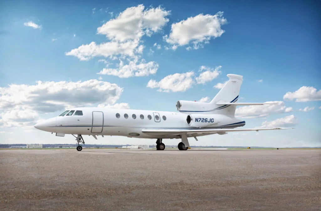 A white Dassault Falcon 900EX business jet with blue stripes, tail number N726JG, parked on an airfield under a blue sky with clouds.