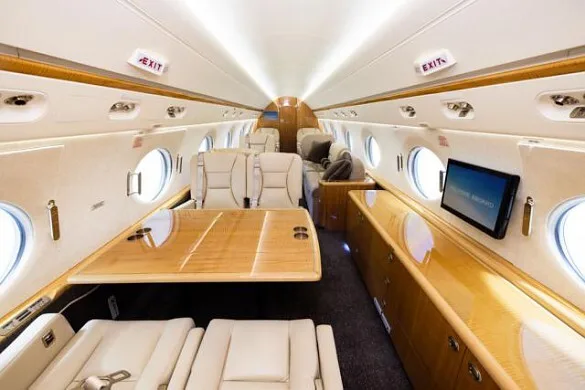 A wide view of a private jet interior featuring a large wooden dining table, beige seats, and a wall-mounted screen.
