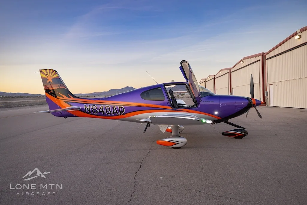 Side view of a purple and silver Cirrus SR22T with open doors, featuring an Arizona flag tail design, parked in front of hangars at sunset.