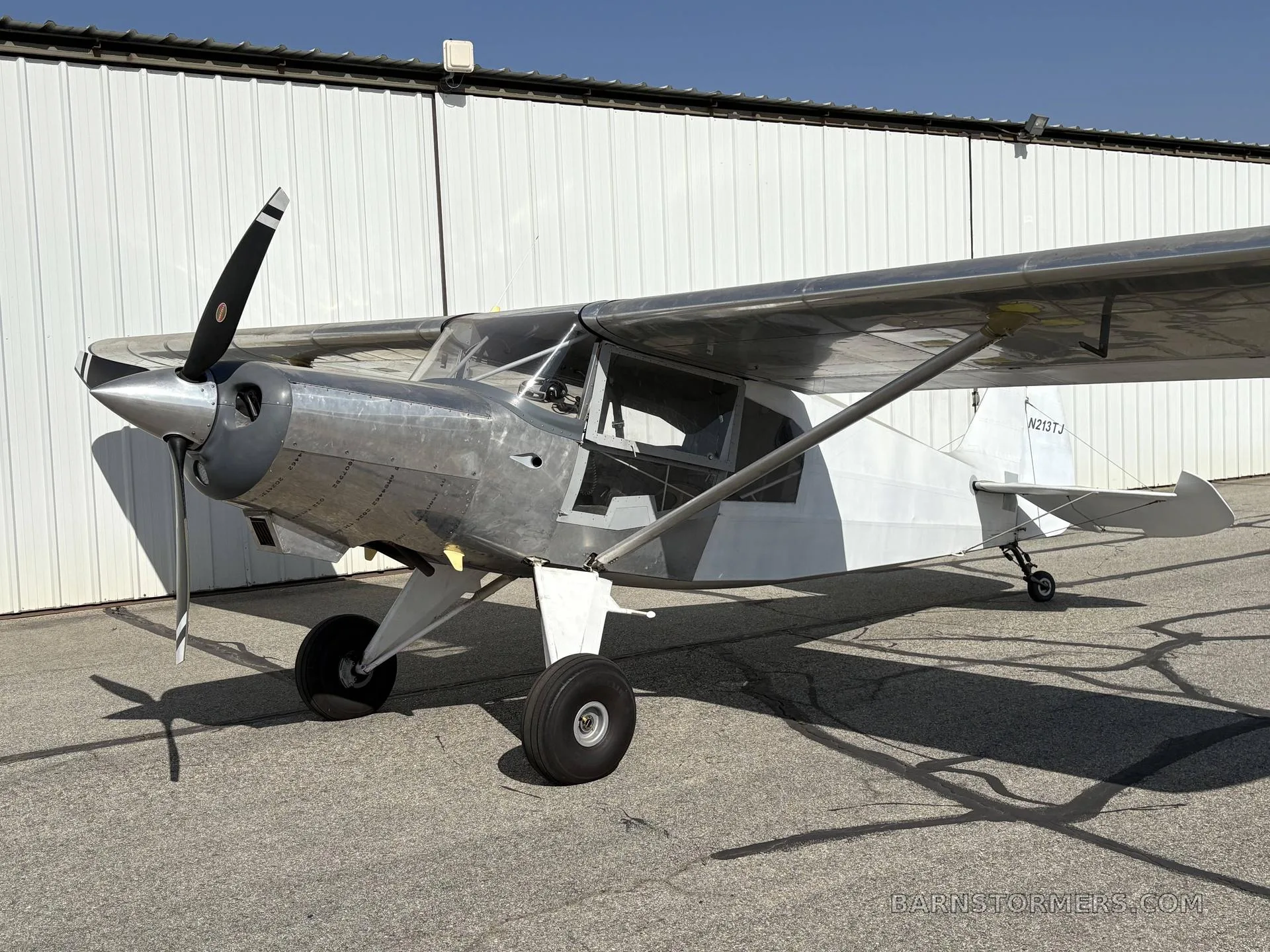 A silver and white single-engine aircraft with a propeller, registration N213TJ, parked on an asphalt tarmac in front of a white hangar.