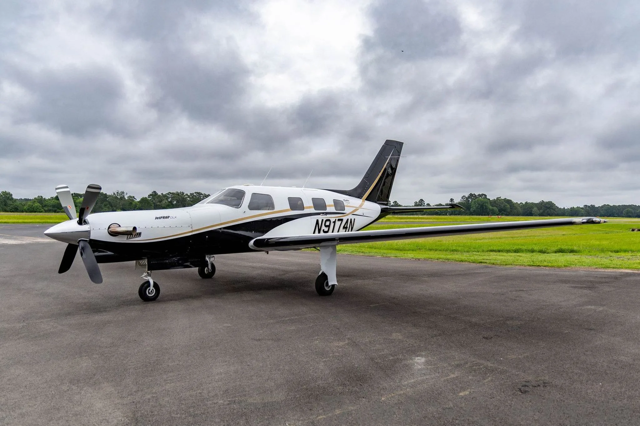 A white and black Piper Malibu Mirage (JetPROP DLX) N9174N with gold accents parked on a tarmac with a grassy field in the background.