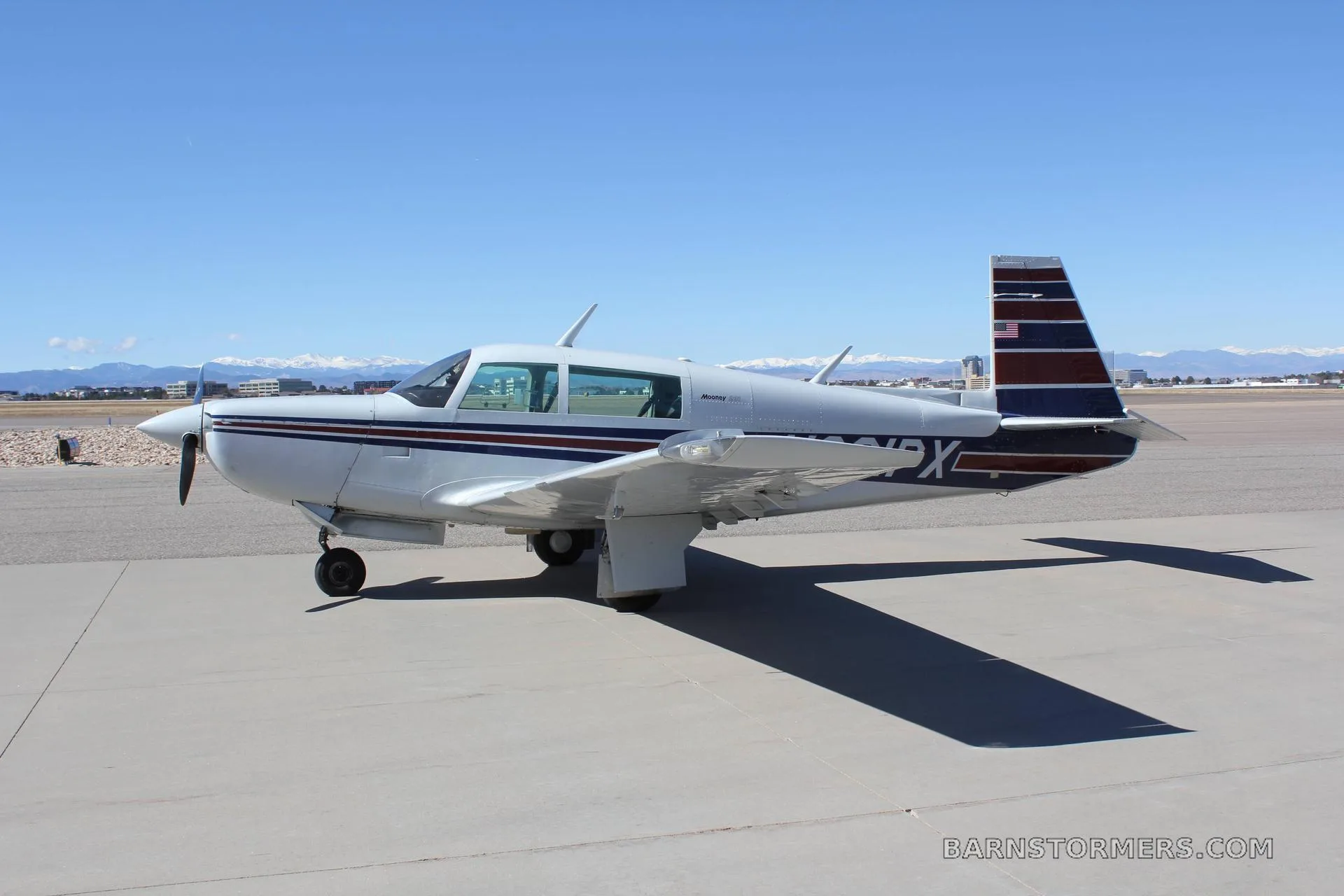 A silver Mooney M20J aircraft with blue and red stripes on the tail and fuselage, parked on an asphalt ramp with mountains in the background.