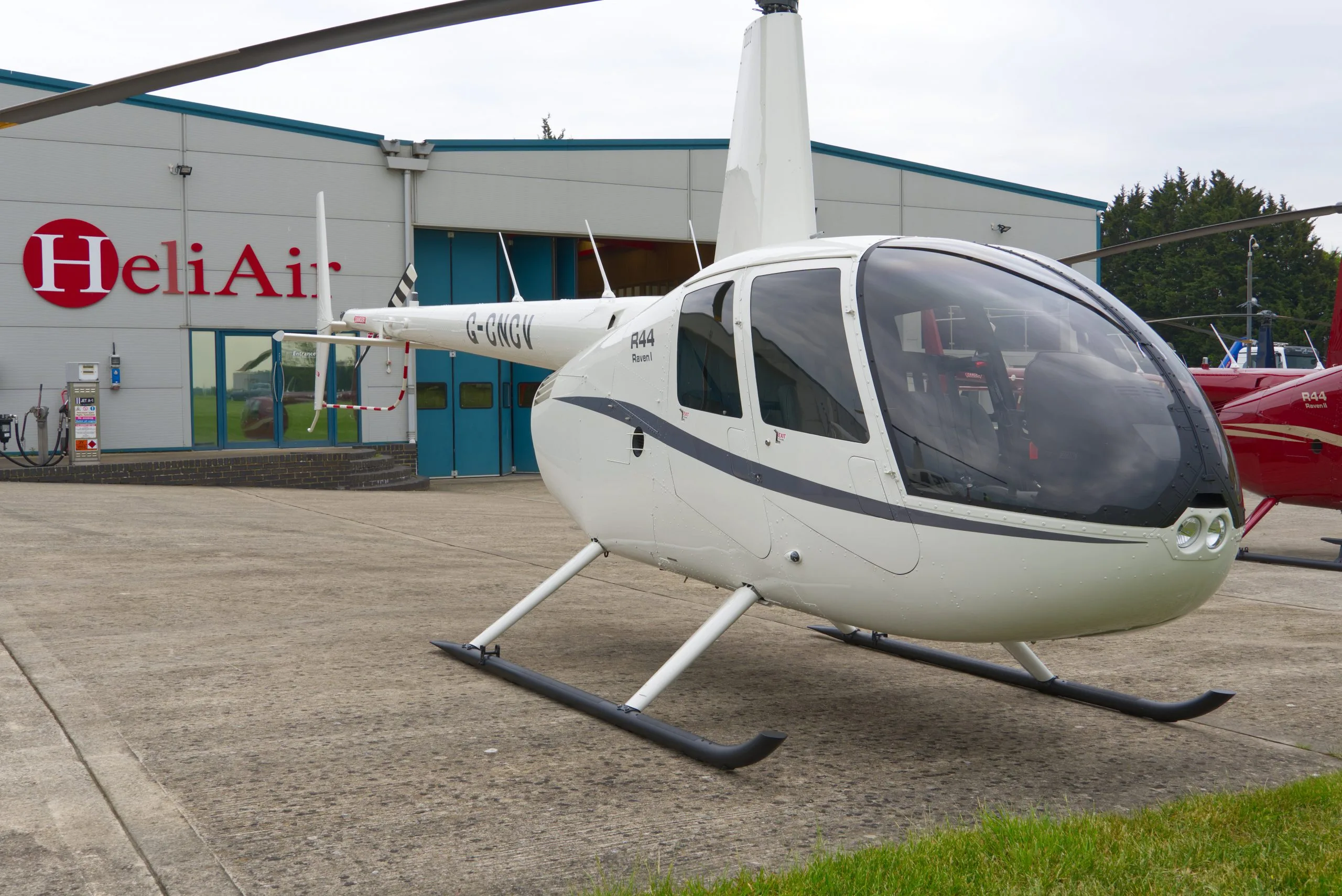 A white Robinson R44 Raven I helicopter (G-GWGV) with dark blue stripes parked on a tarmac in front of a HeliAir hangar.