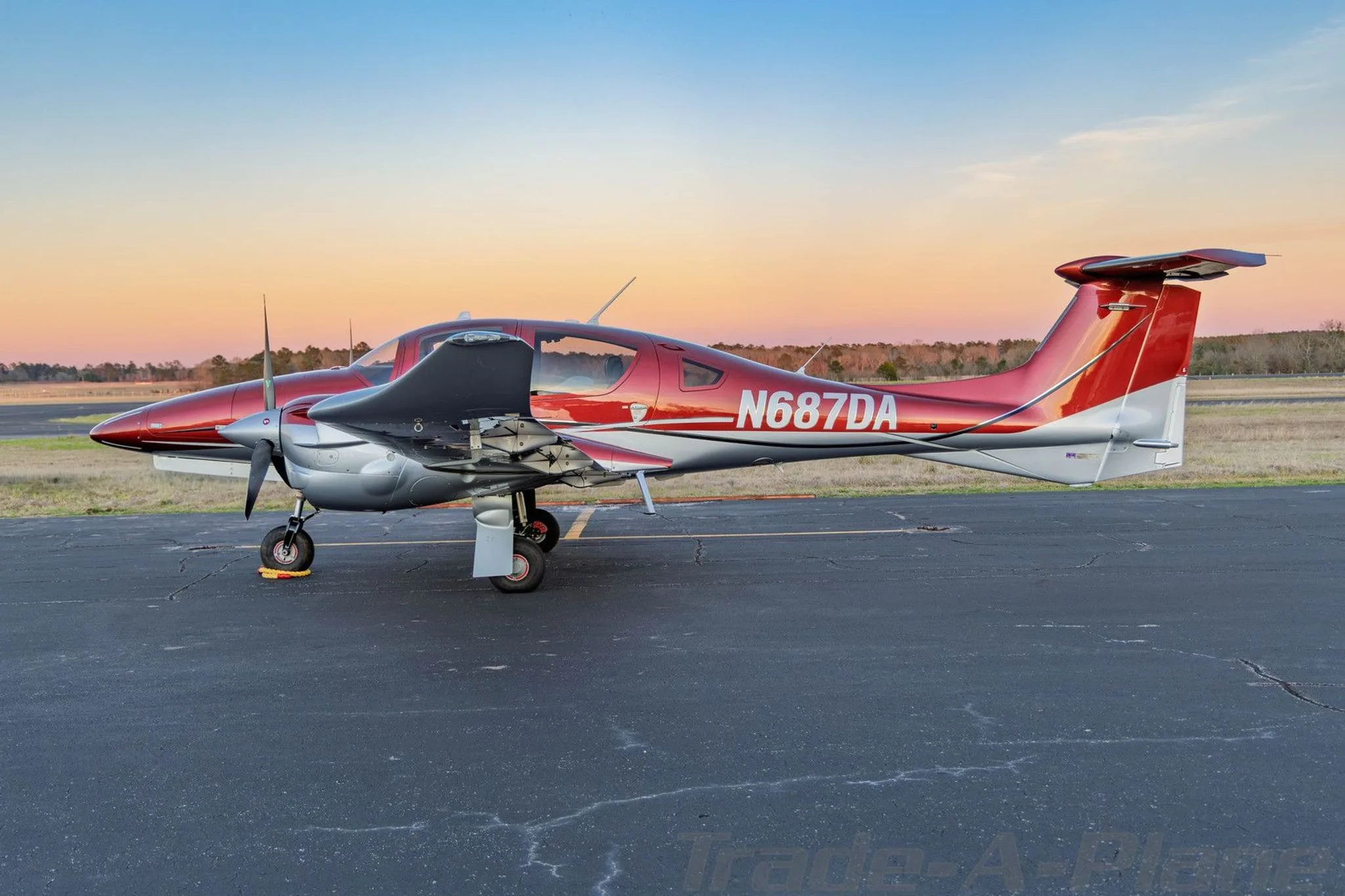 A three-quarter rear view of a red and silver Diamond DA62 aircraft parked on an airfield at sunset.
