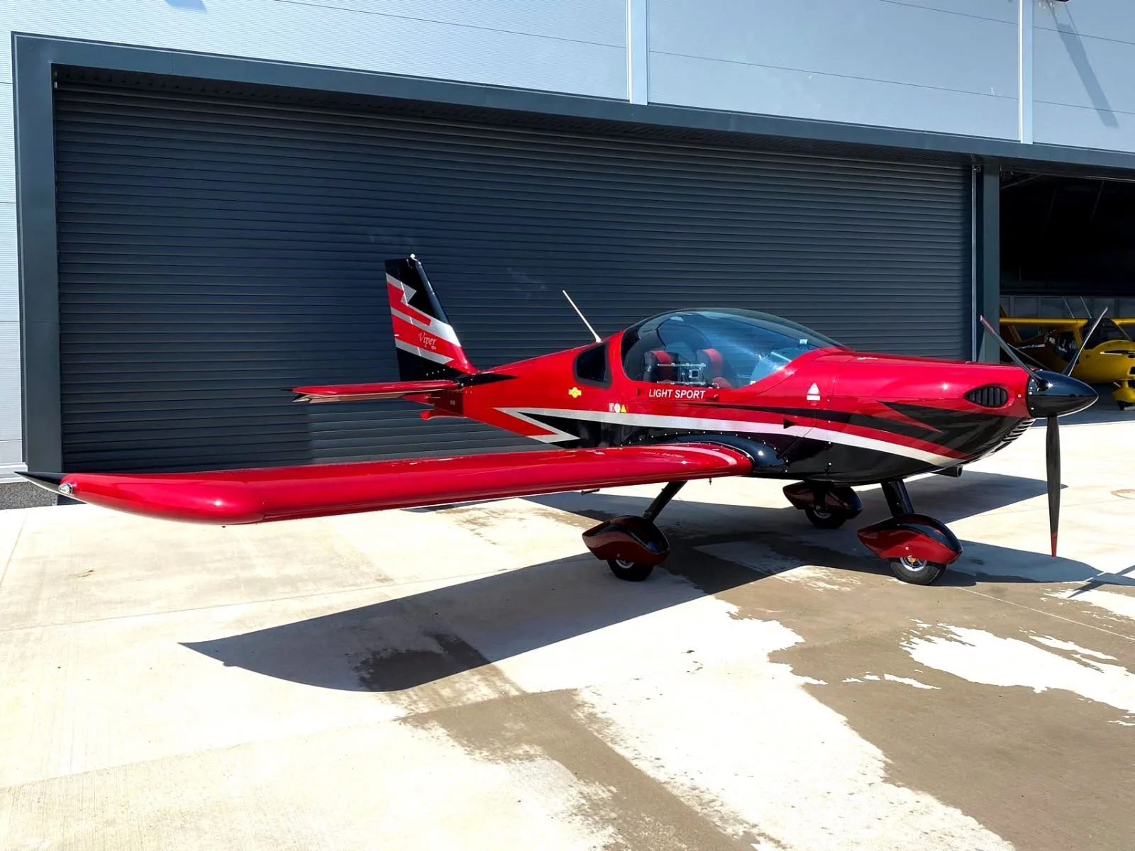 A red and black Viper SD4 light sport aircraft with its canopy closed, parked on a concrete apron next to a hangar.