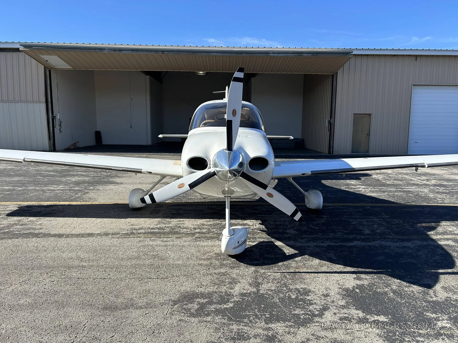 A white Cirrus SR22 aircraft with a three-bladed propeller, registration N948CD, viewed head-on in front of an open hangar.