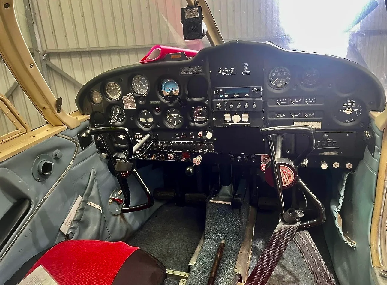 The cockpit of a light aircraft features a black instrument panel with various gauges and controls, and a red seat cushion.