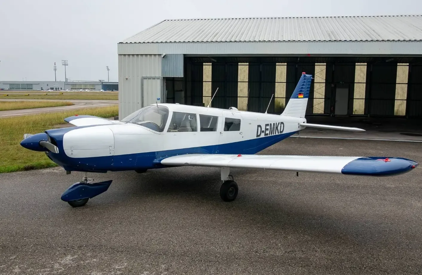 A white and blue single-engine propeller aircraft (D-EMKD) parked on an asphalt apron next to a hangar.