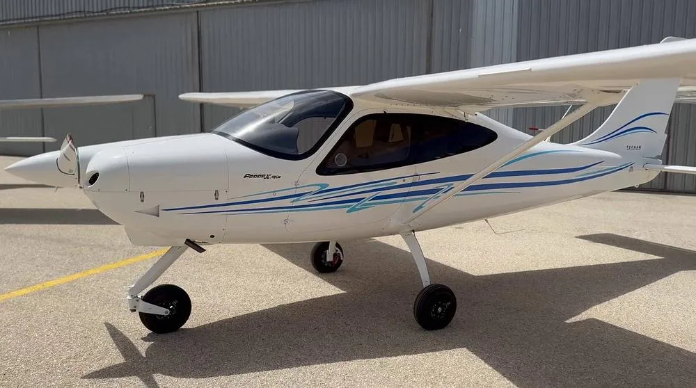 A close-up side view of a white Tecnam P2008 aircraft with blue and light blue stripes, parked on an airfield.