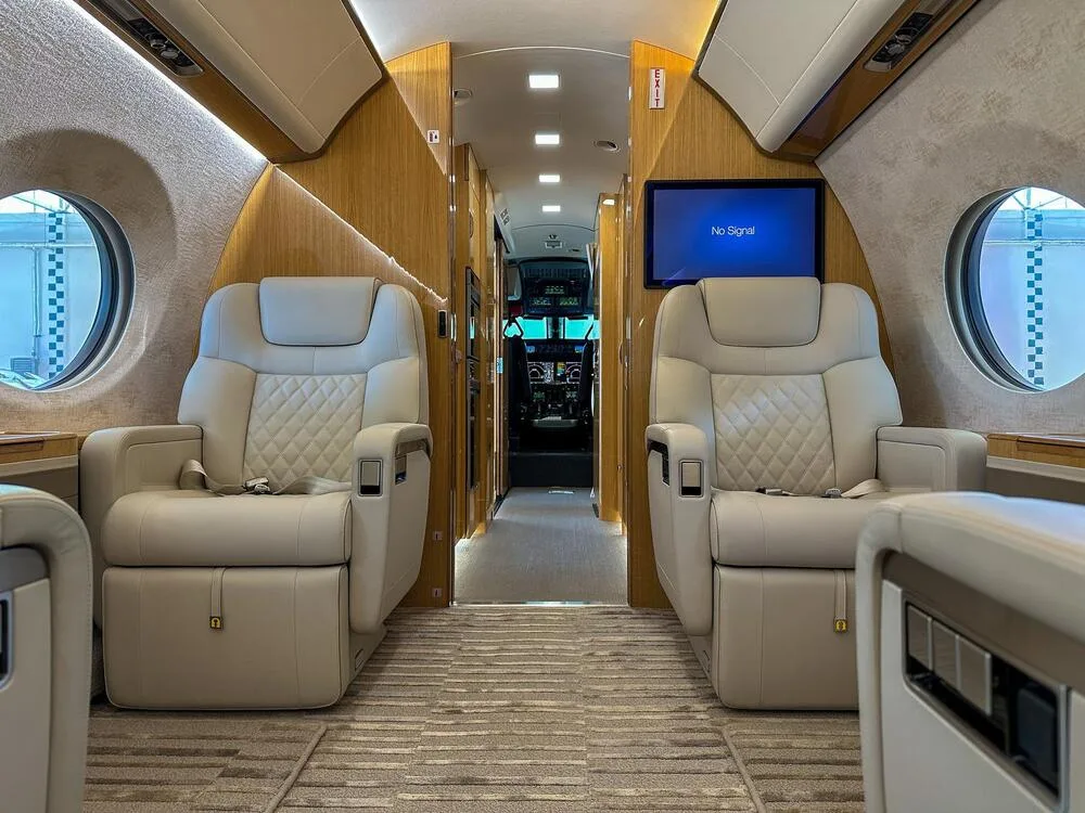 View down the aisle of a luxury aircraft cabin with light-colored quilted leather seats and wood paneling, looking towards the cockpit.