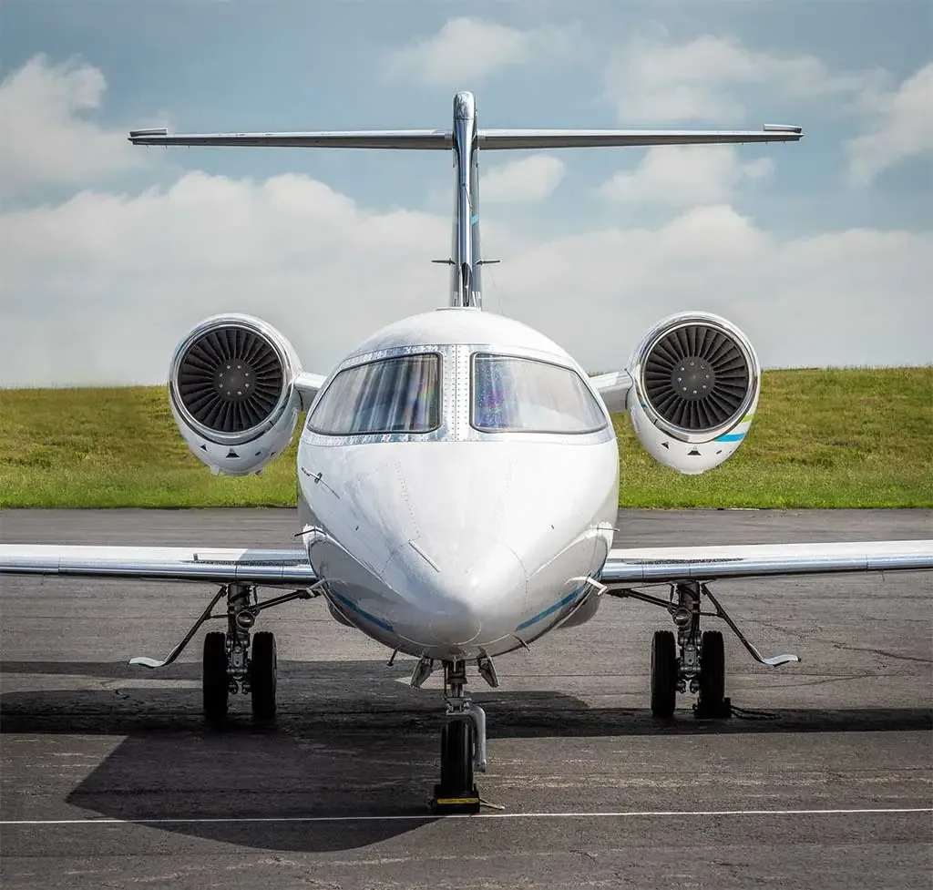 A head-on view of a white business jet with blue and green stripes, parked on an asphalt tarmac under a partly cloudy sky.