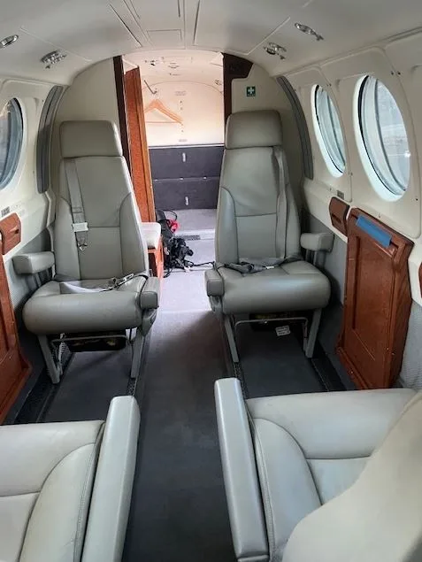 The interior of an aircraft cabin with two rows of light grey leather seats facing each other, and a view towards the rear.