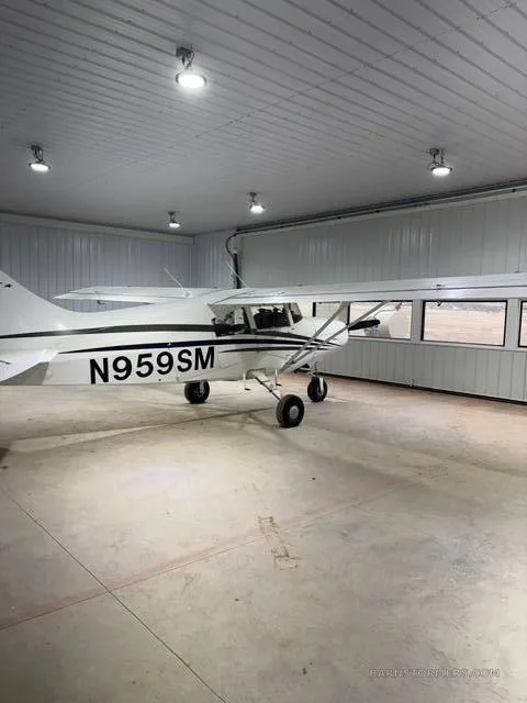 A white Maule M-7-235B aircraft with black stripes and registration N959SM is parked inside a hangar, viewed from the side.