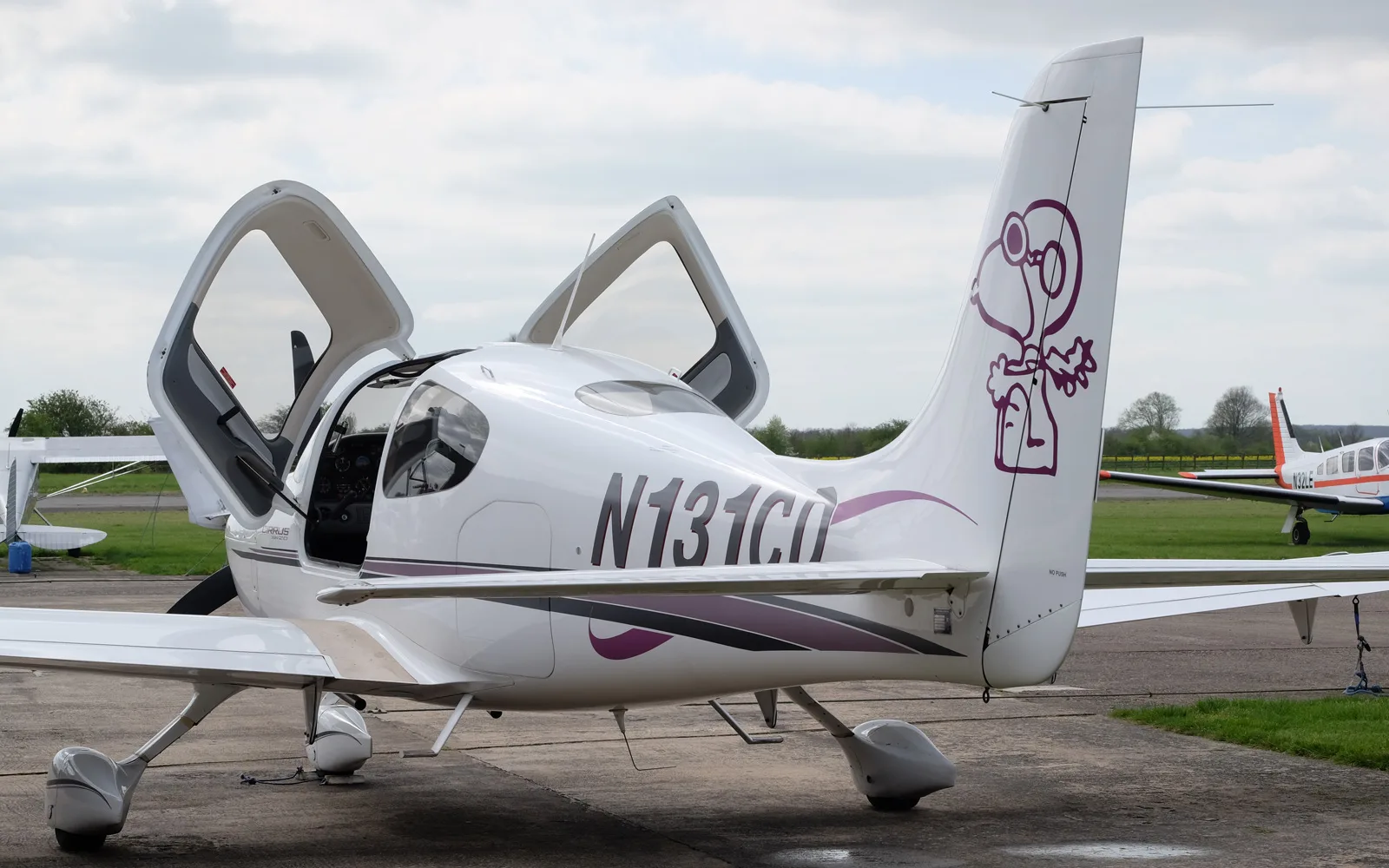 A rear view of a white Cirrus SR22 aircraft with open gull-wing doors and a prominent Snoopy graphic on the tail.