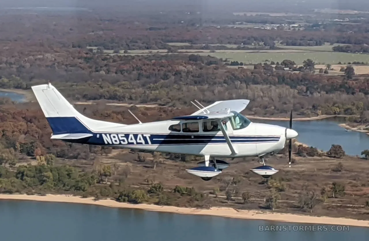 A white and blue Cessna 182 aircraft flies low over a lake and autumn trees.