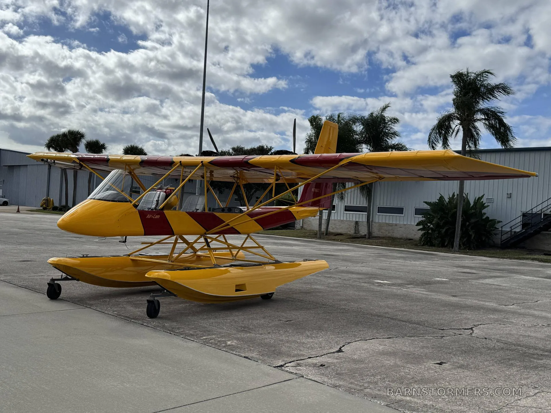 A yellow and red Air-Cam seaplane with floats, viewed from the front-left side, parked on an asphalt ramp with buildings and palm trees in the background.