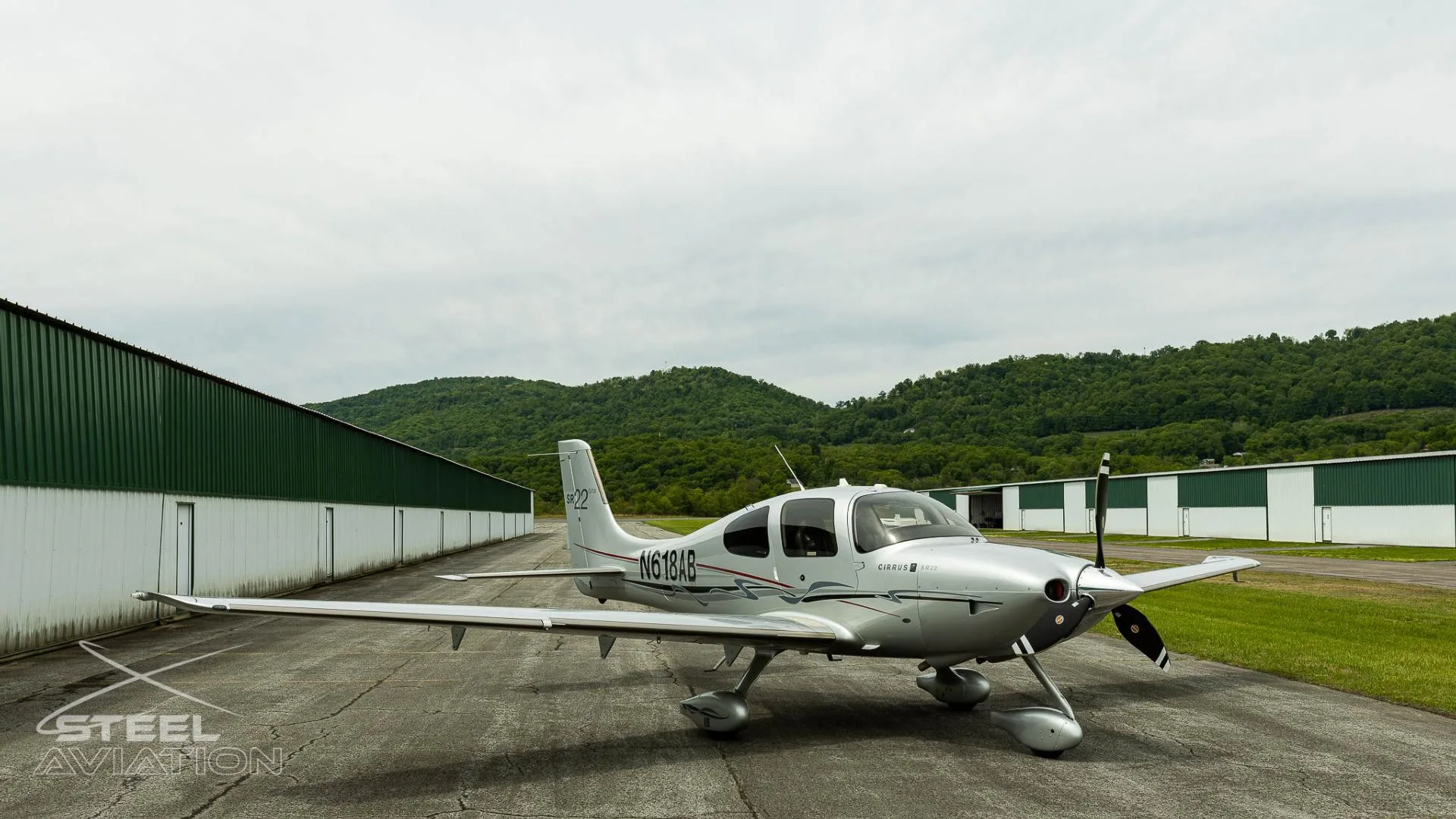 A silver Cirrus SR22 GTS aircraft with registration N618AB parked on an tarmac with hangars and green hills in the background.