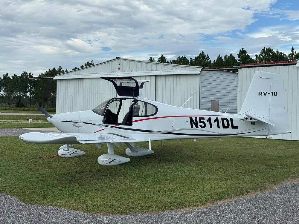 A white Van's RV-10 aircraft with red and black accents, registration N511DL, parked on grass with its gull-wing doors open, next to a hangar.