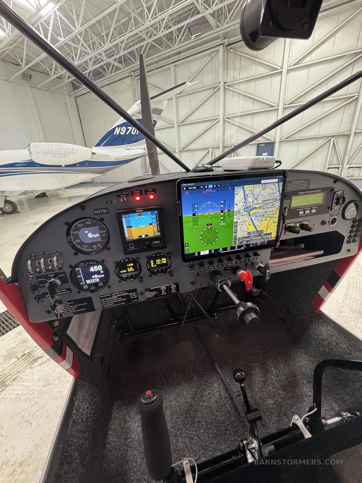 A cockpit view of the instrument panel with multiple screens, inside a hangar, with another aircraft visible through the windshield.