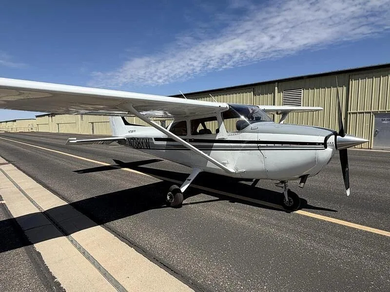 A front-right view of the white Cessna 172, N738YW, with gray and black stripes, parked on a tarmac next to a hangar.
