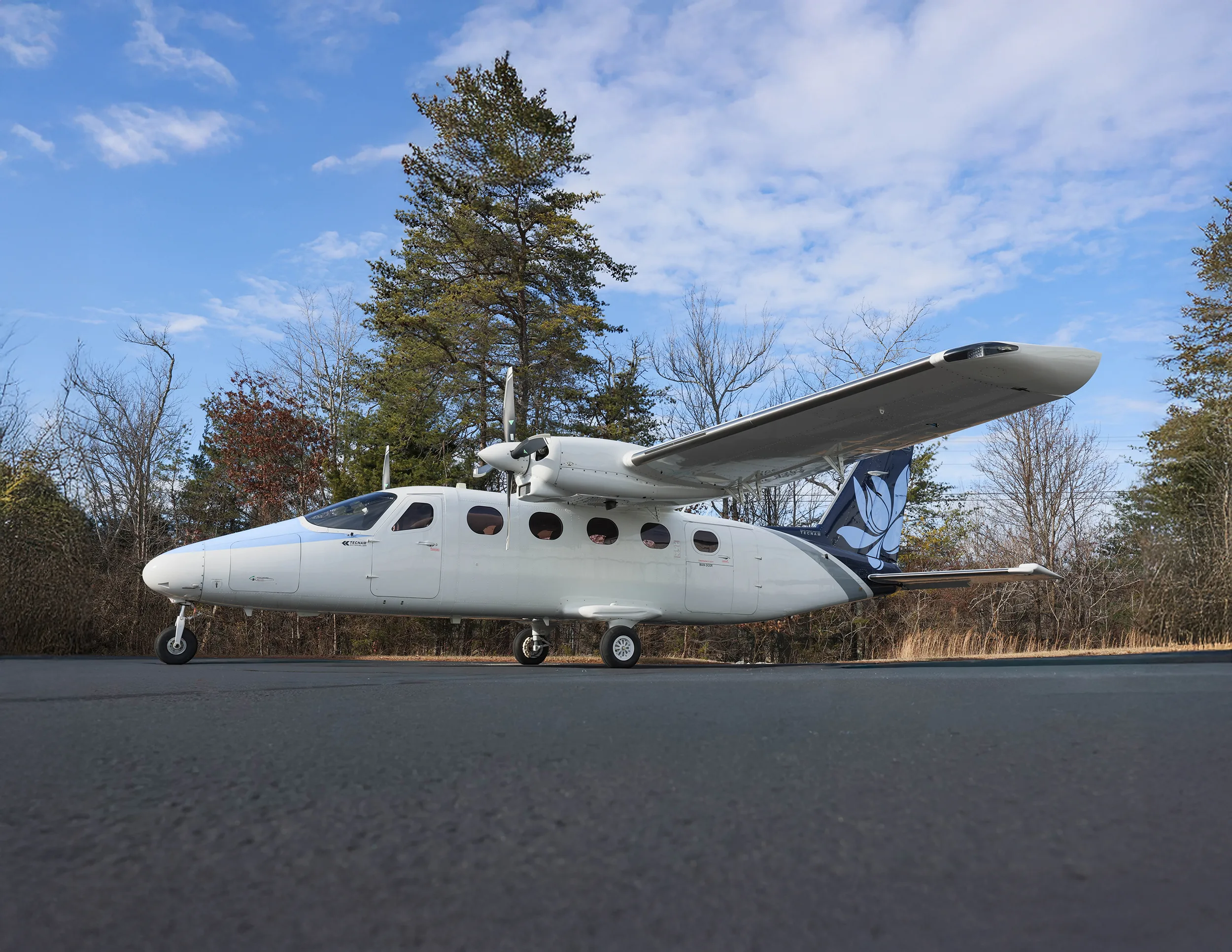 A white and blue twin-engine aircraft with a floral tail logo parked on an asphalt ramp with trees in the background.