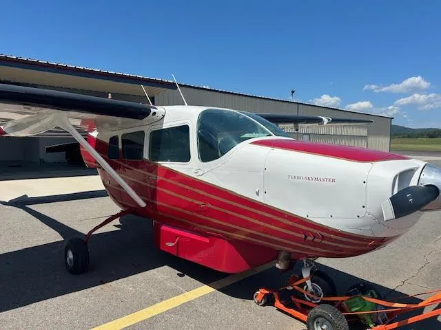 A white and red Cessna Turbo Skymaster aircraft is parked on an asphalt tarmac with a hangar in the background.