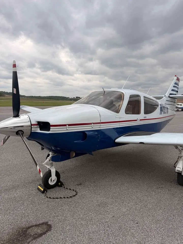 A front-left view of a white and blue single-engine aircraft with registration N4921W on the tarmac under a cloudy sky.