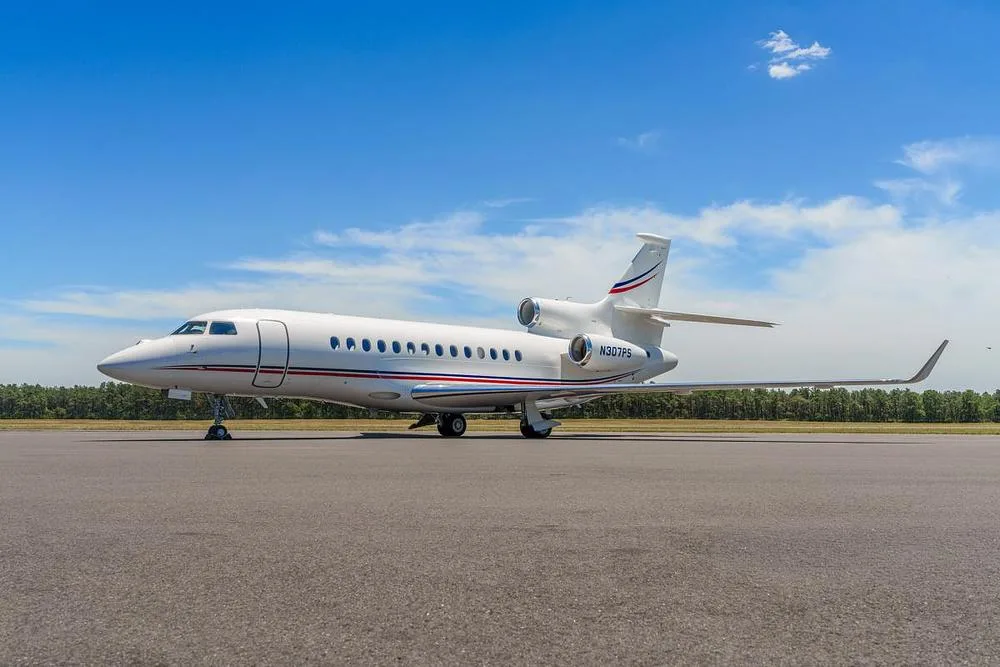 A white Dassault Falcon 900EX business jet with red and blue stripes on the tail and fuselage, parked on an asphalt runway under a clear blue sky.