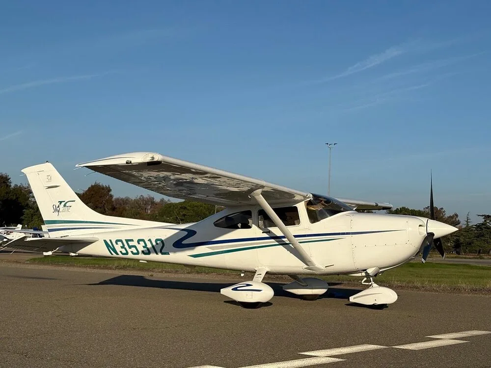 A white Cessna 182 Skylane with blue and green stripes, registration N35312, parked on an tarmac under a clear blue sky.