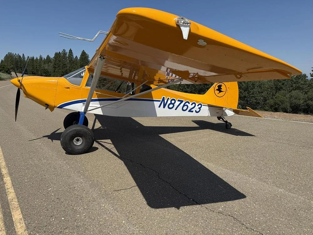 A yellow and white high-wing aircraft, N87623, with large tires, is parked on an asphalt runway under a clear blue sky.
