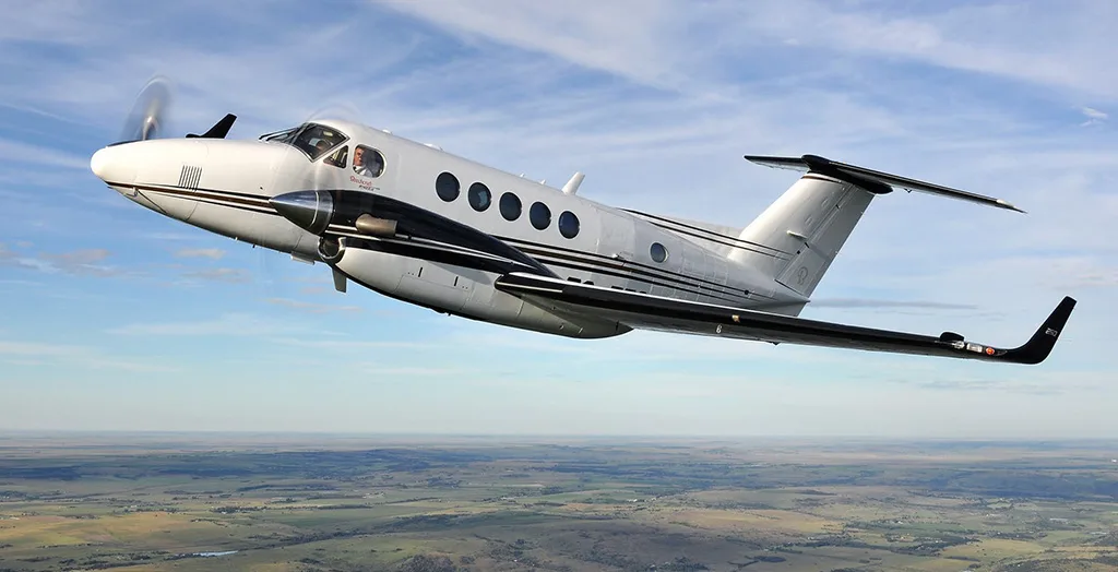 A white and black Beechcraft King Air 250 turboprop aircraft flying over a green and brown landscape under a partly cloudy sky.