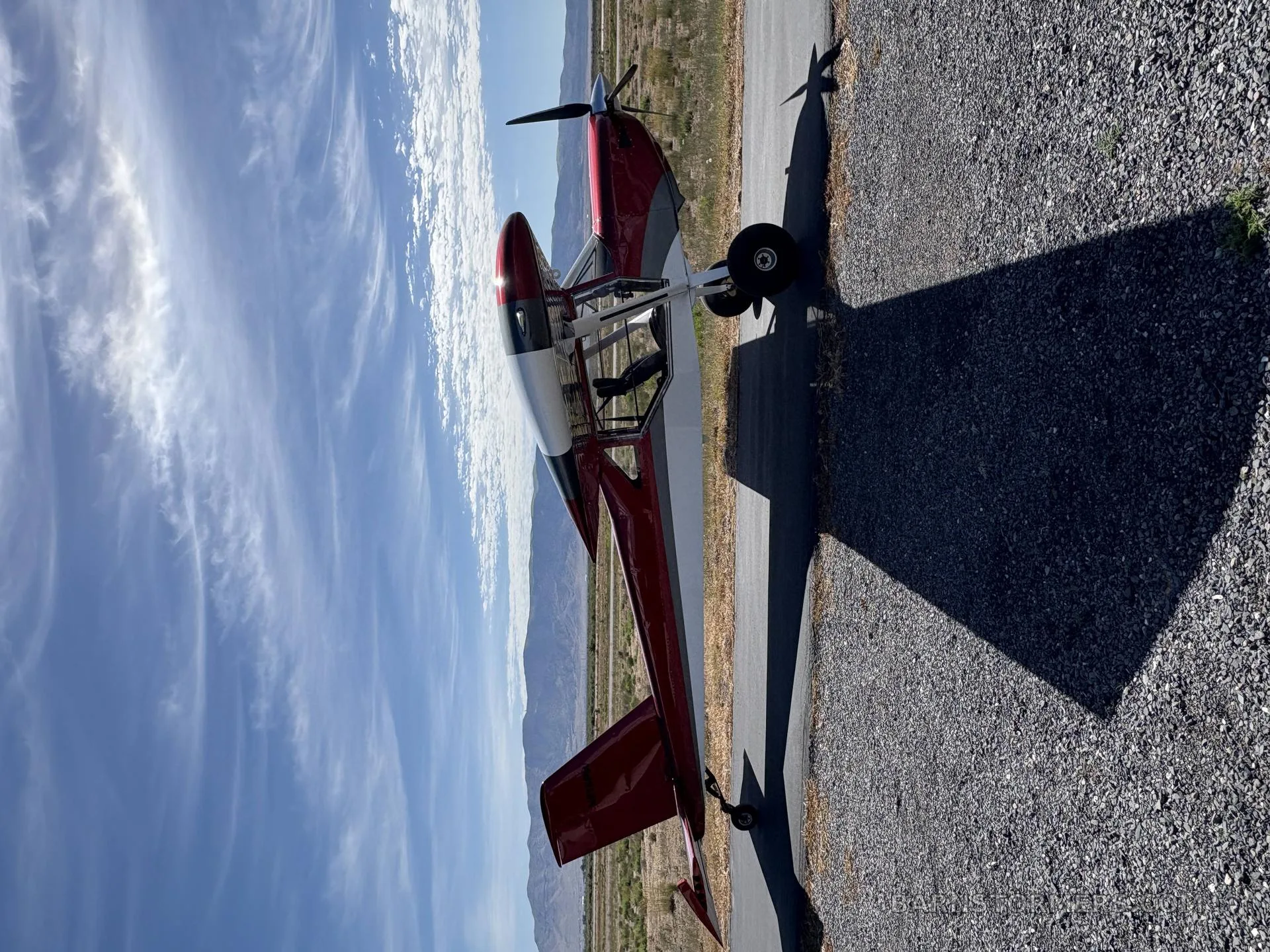 A red and white single-engine aircraft with a propeller on an asphalt strip, viewed from the side with mountains in the background.