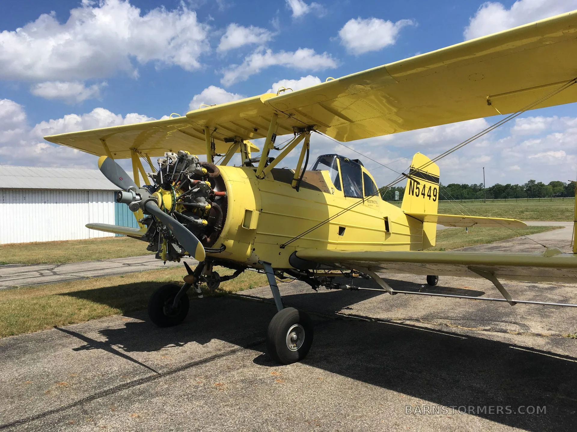 A yellow biplane crop duster with its radial engine exposed, parked on an asphalt ramp under a partly cloudy sky.