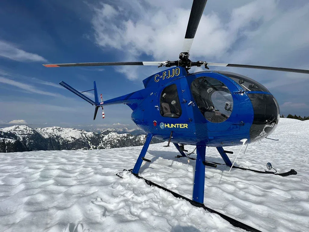 A blue helicopter with registration C-FJJ0 sits on a snowy mountain peak under a blue sky.
