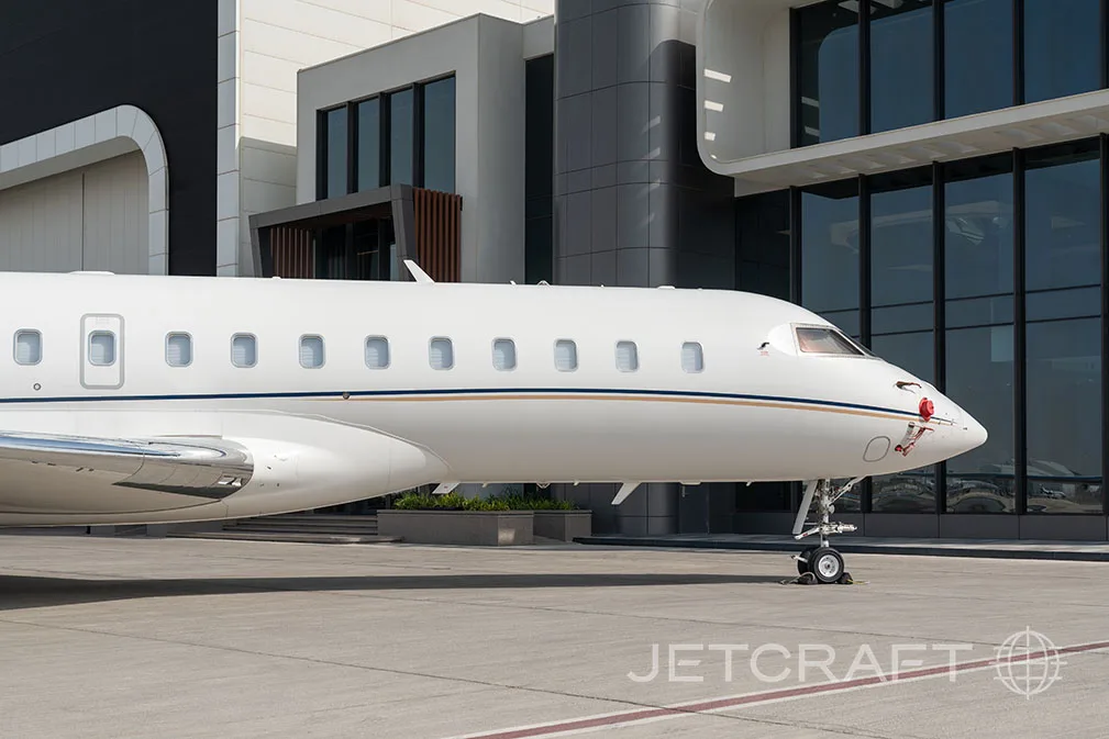A white business jet with a gold and blue stripe parked on a tarmac in front of a modern building, showing the front fuselage and nose.