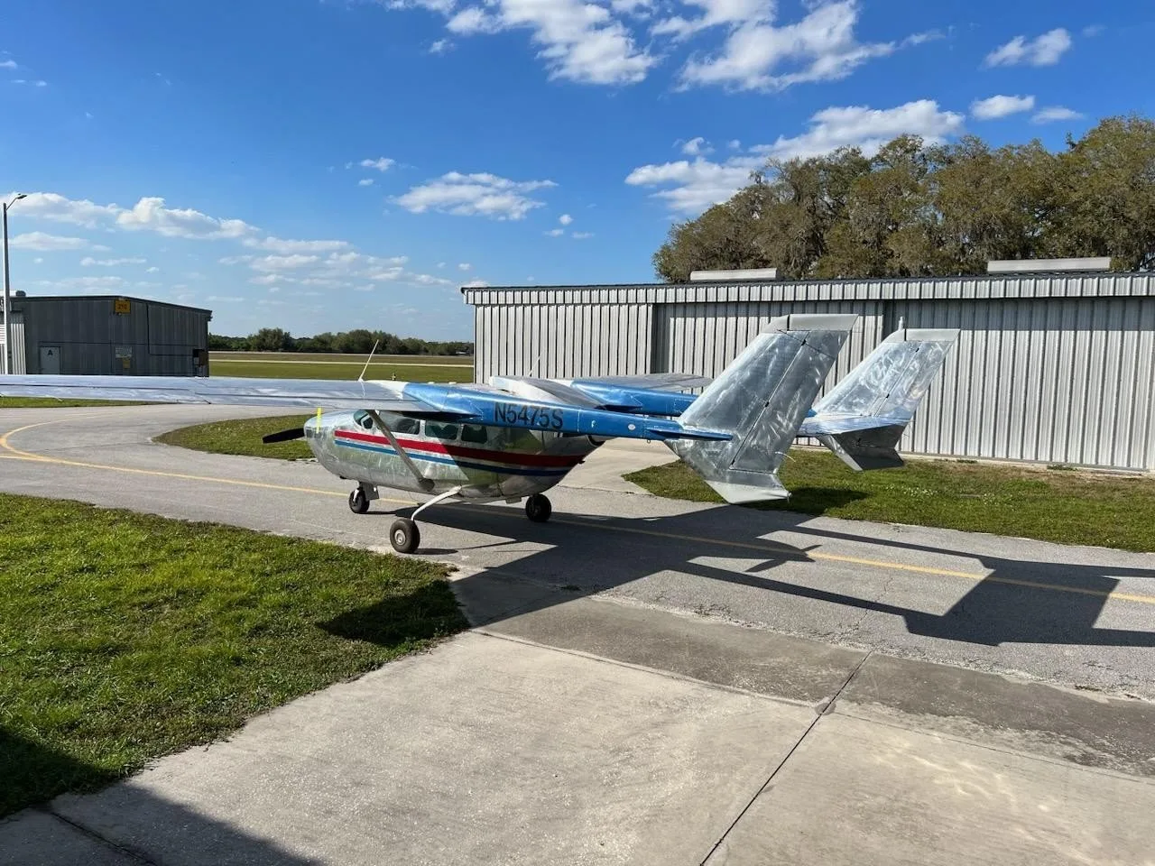 A silver and blue single-engine aircraft with twin tail booms, registration N5475S, parked on an airfield next to a hangar.