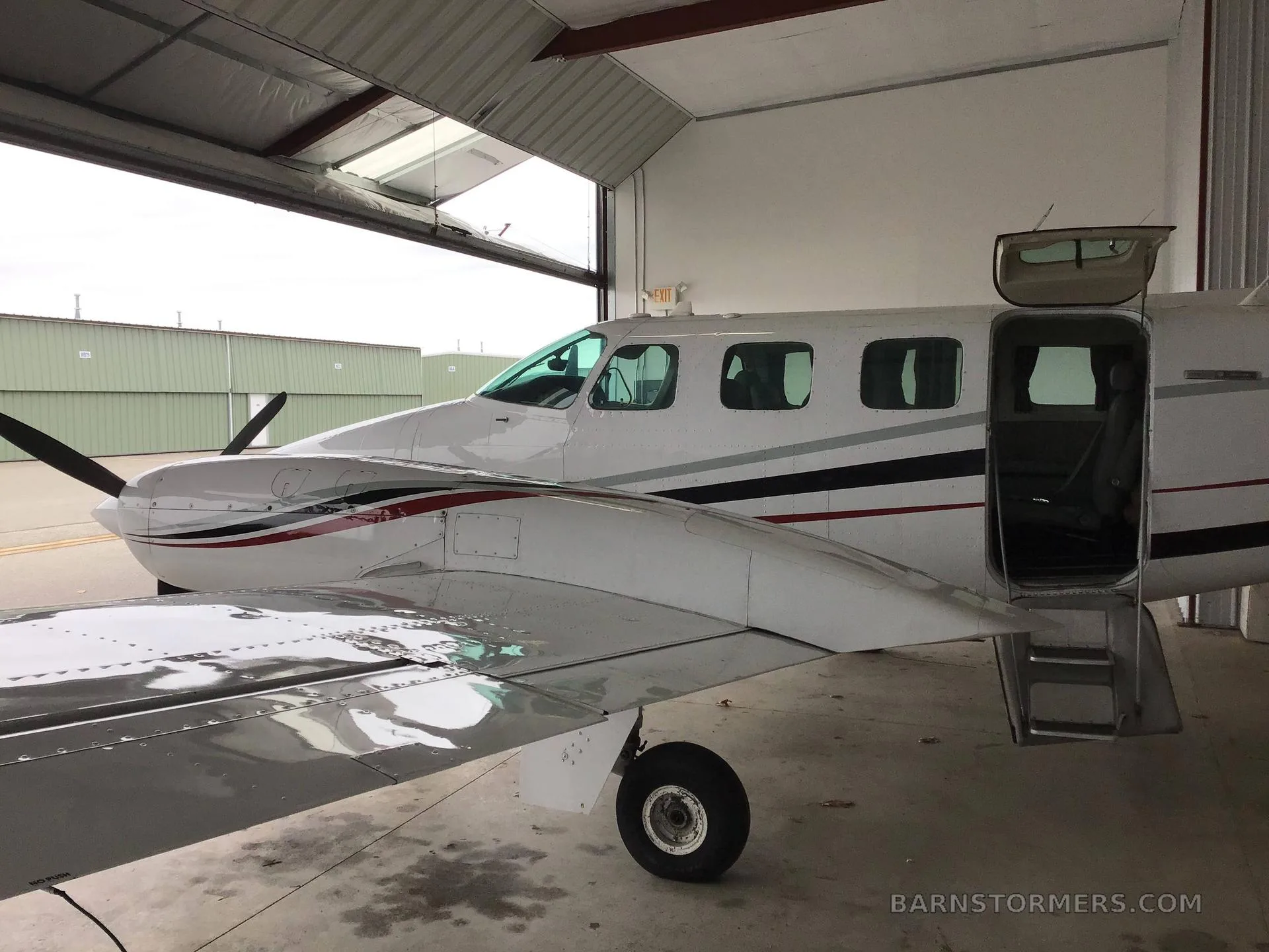 A white twin-engine propeller aircraft with red and black stripes, passenger door open, partially parked in a hangar with the door open to the outside.